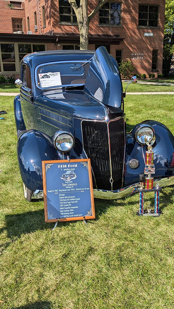 Front three-quarter view of a dark blue 1936 Ford coupe on grass with an open hood, framed build specification board, and award trophy.