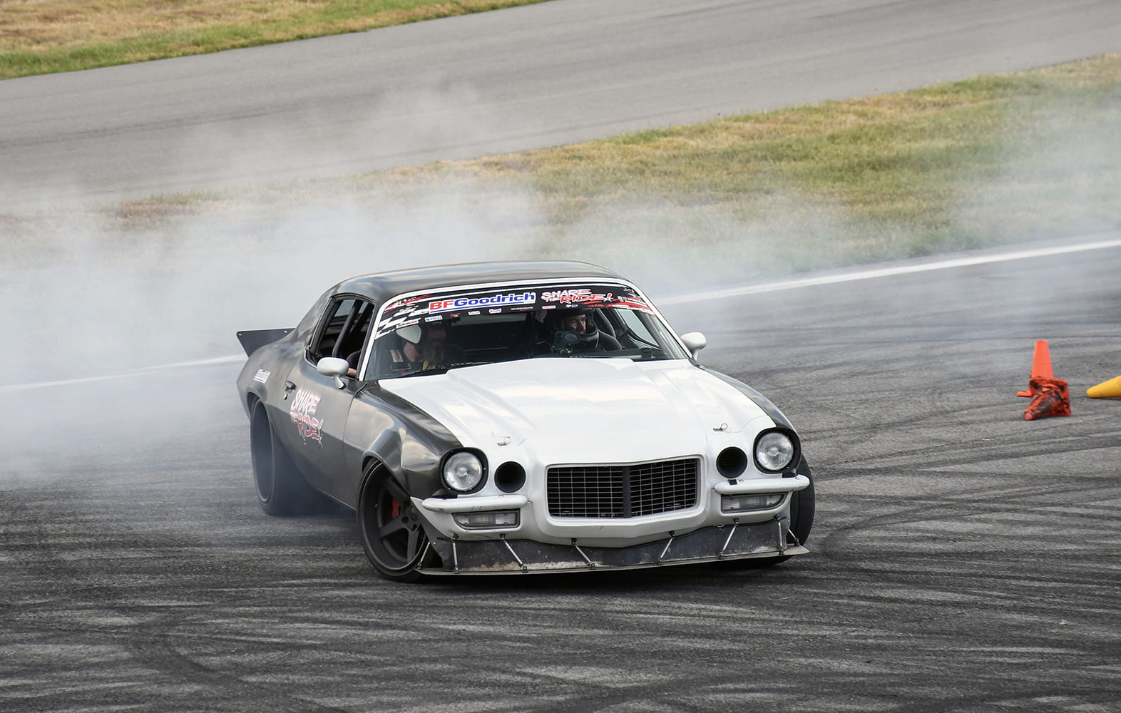 Black and white second-generation Chevrolet Camaro drifting through a turn with tire smoke and orange cones on an autocross course.