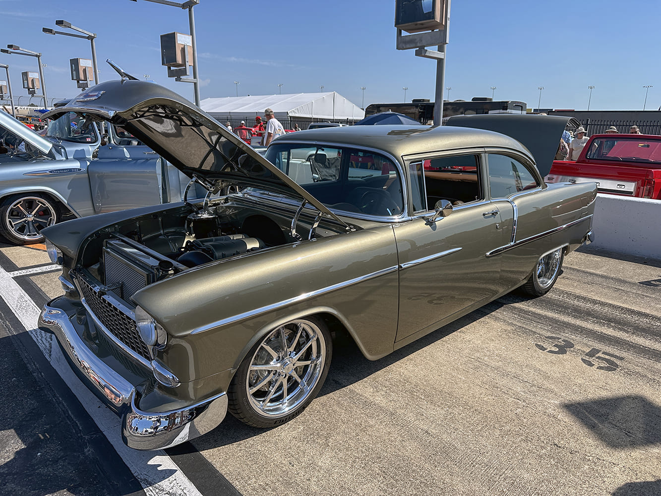 Front three-quarter angle of an olive-bronze metallic 1955 Chevrolet two-door sedan with modern chrome wheels and open hood on a speedway pit lane.