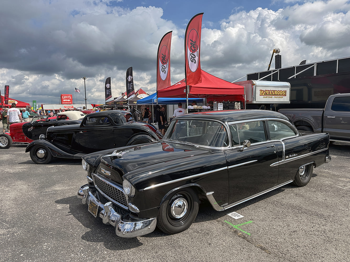 Classic gloss black 1955 Chevrolet Bel Air two-door post sedan with black steel wheels and chrome hubcaps displayed under event flags.