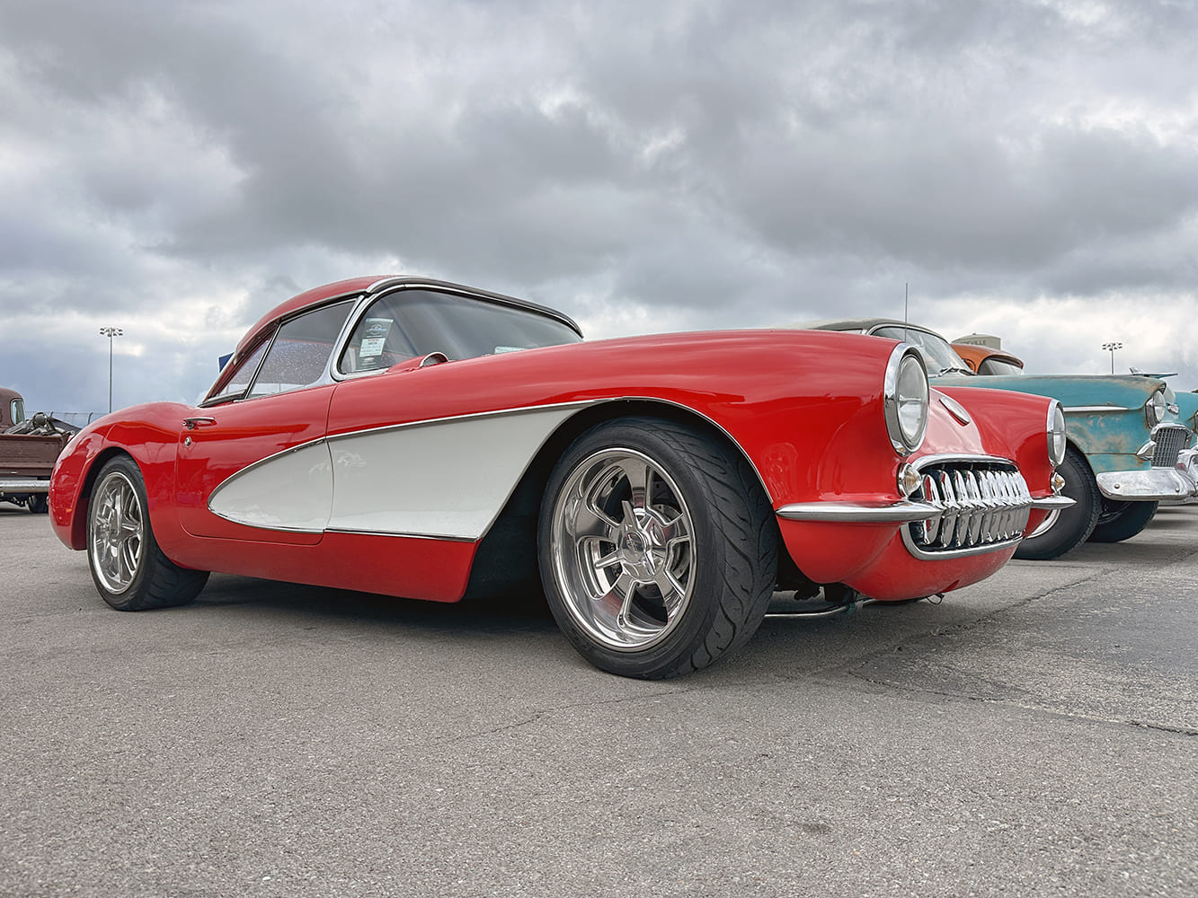 Low front three-quarter profile of a red 1957 Chevrolet Corvette roadster with white coves, tooth grille, and modern polished five-spoke wheels.