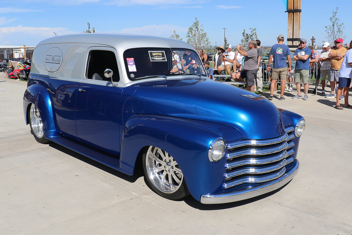 Front three-quarter view of a lowered two-tone blue and silver vintage Chevrolet panel delivery truck rolling past spectators seated along the event perimeter.