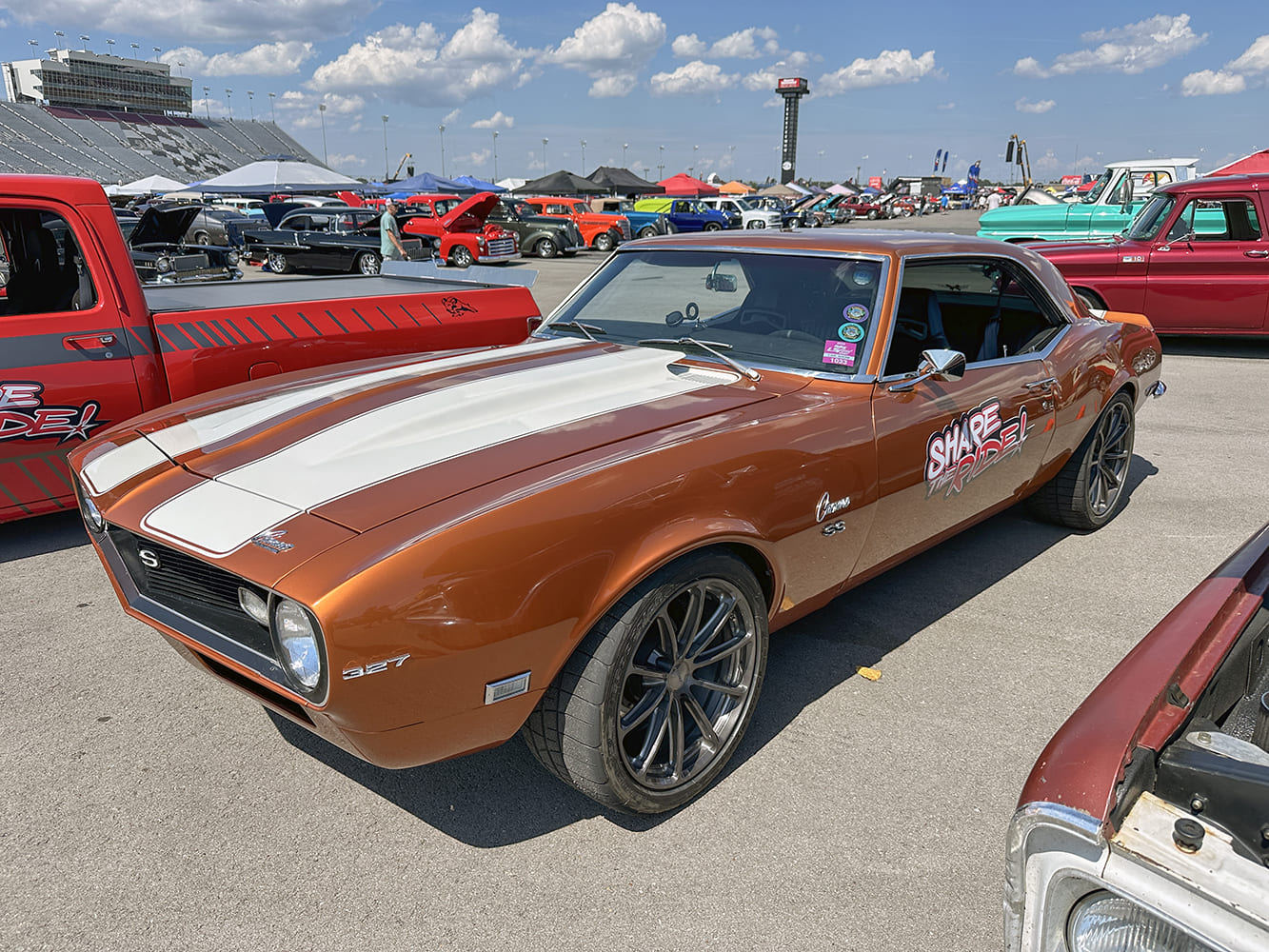Front three-quarter view of a metallic copper-bronze 1968 Chevrolet Camaro with white hood stripes, modern charcoal wheels, and Share The Ride door graphics.