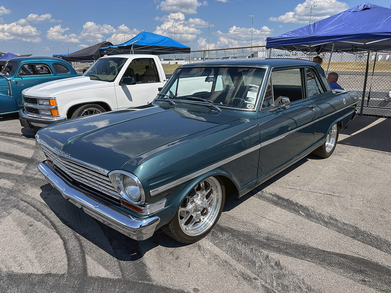 High front three-quarter angle of a dark teal metallic 1964 Chevy II Nova two-door sedan on modern chrome billet wheels at an outdoor car meet.