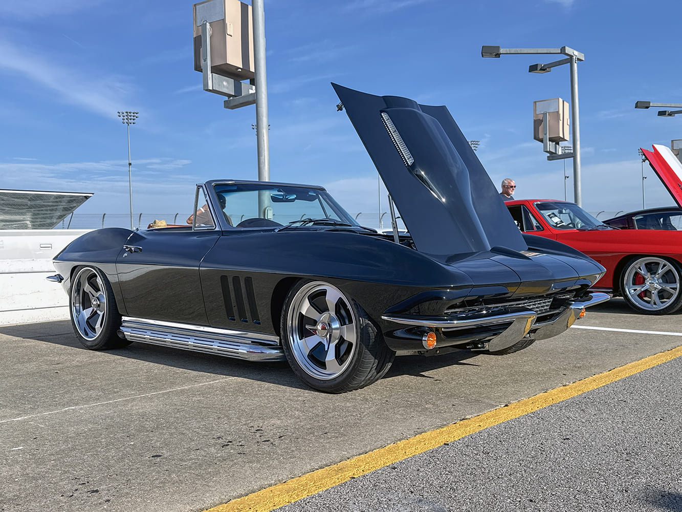 Low side three-quarter profile of a glossy black 1966 Corvette C2 convertible with side exhaust pipes, modern wheels, and open stinger hood.
