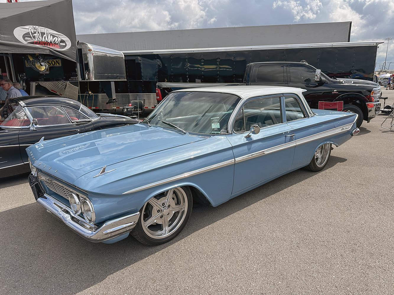 Front three-quarter view of a powder-blue 1961 Chevrolet Biscayne sedan with a white top and modern five-spoke chrome wheels parked by a hauler trailer.
