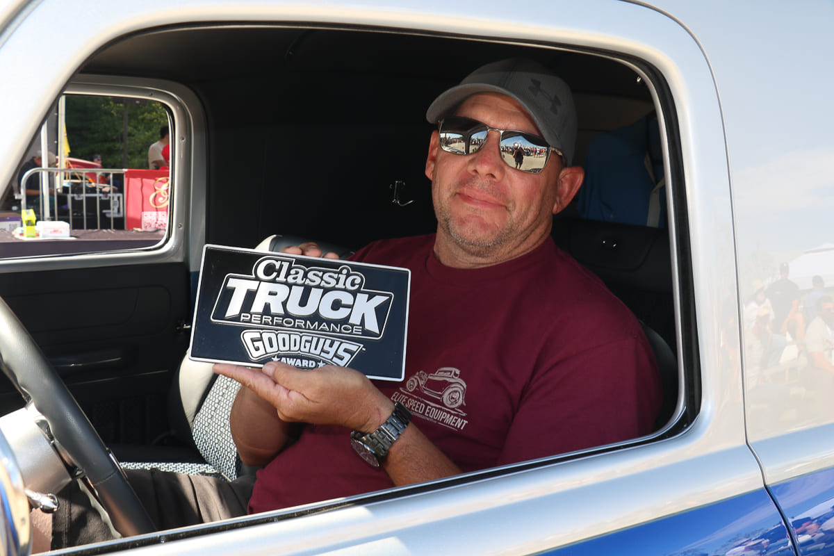 Driver inside a vintage truck holding up the Classic Truck Performance Good Guys Award plaque toward the camera.