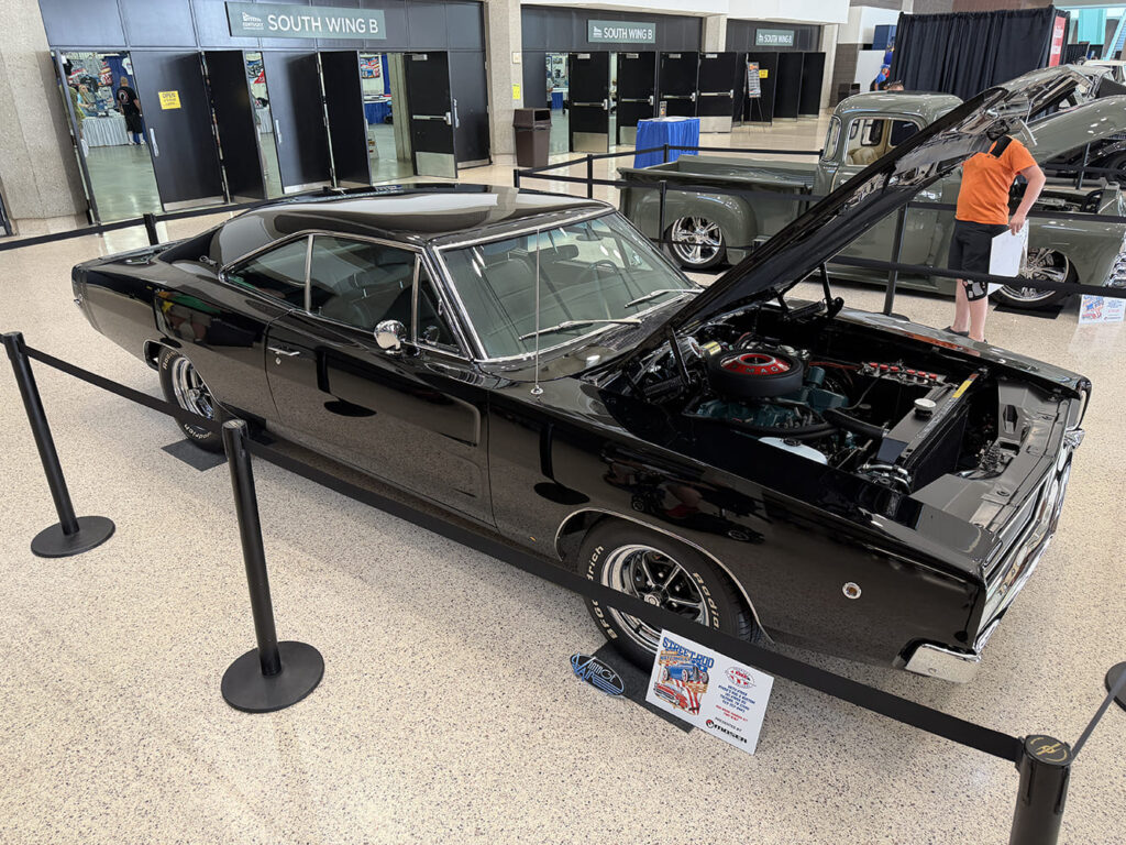 Front three-quarter perspective of a glossy black 1968 Dodge Charger muscle car with open hood, Magnum 500-style wheels, and redline tires inside the convention hall entrance.