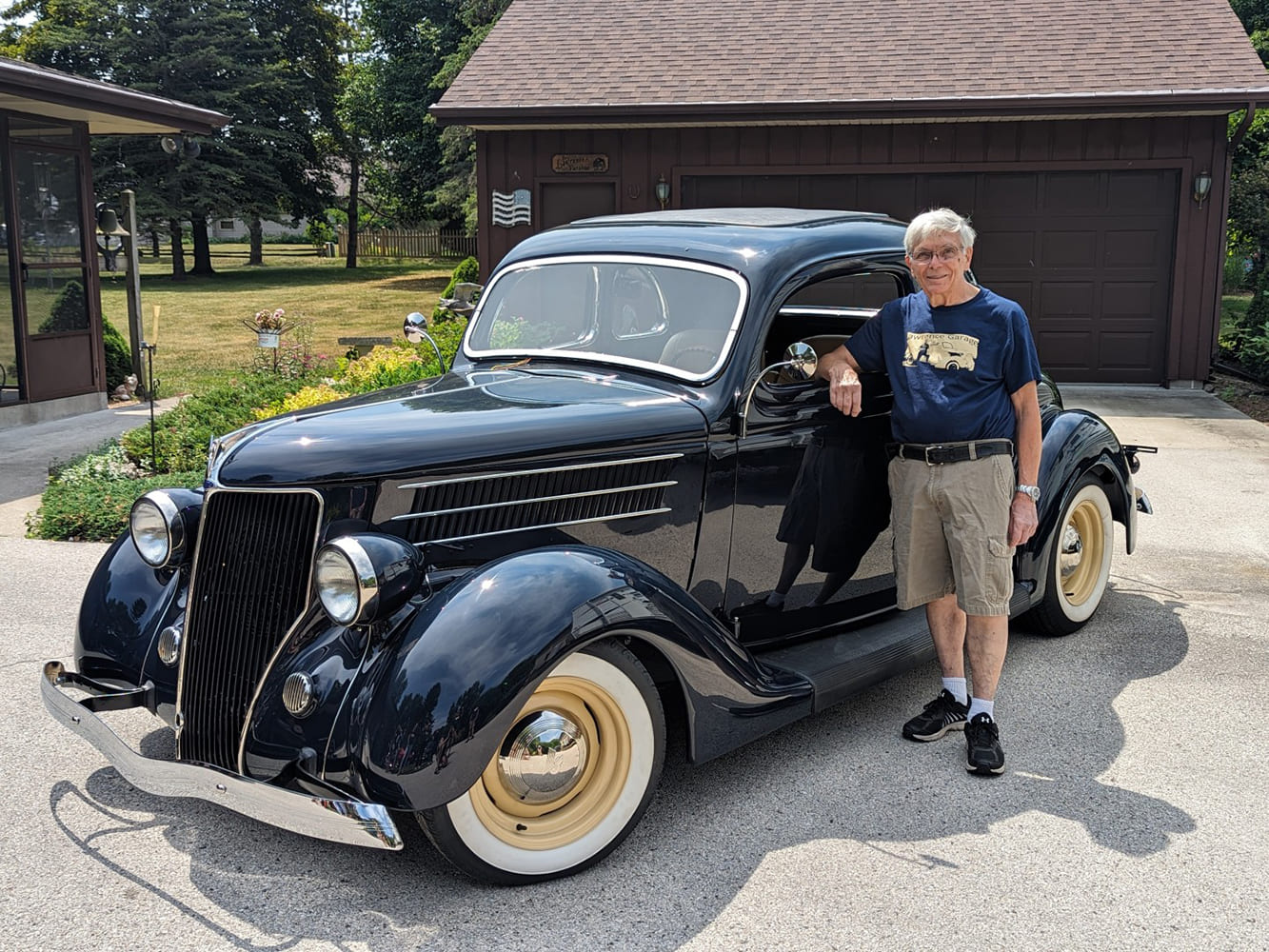 Tom Lawrence leaning against the driver-side door of his dark blue 1936 Ford five-window coupe parked outside a residential garage.