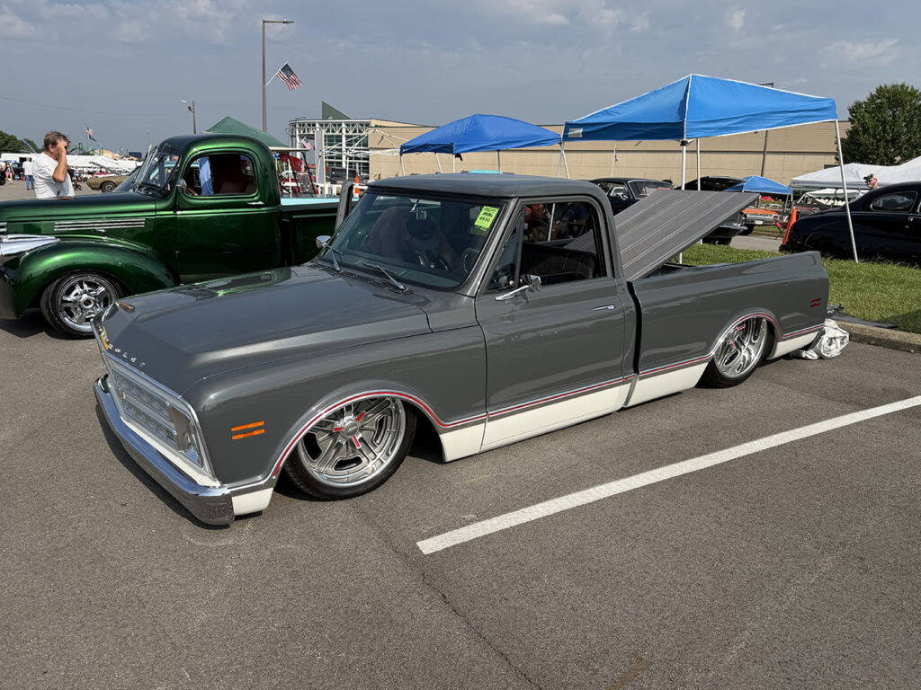 Low front three-quarter profile of a lowered two-tone dark gray and white Chevrolet C10 pickup with a raised hard tonneau cover parked in a sunny outdoor fairgrounds row.