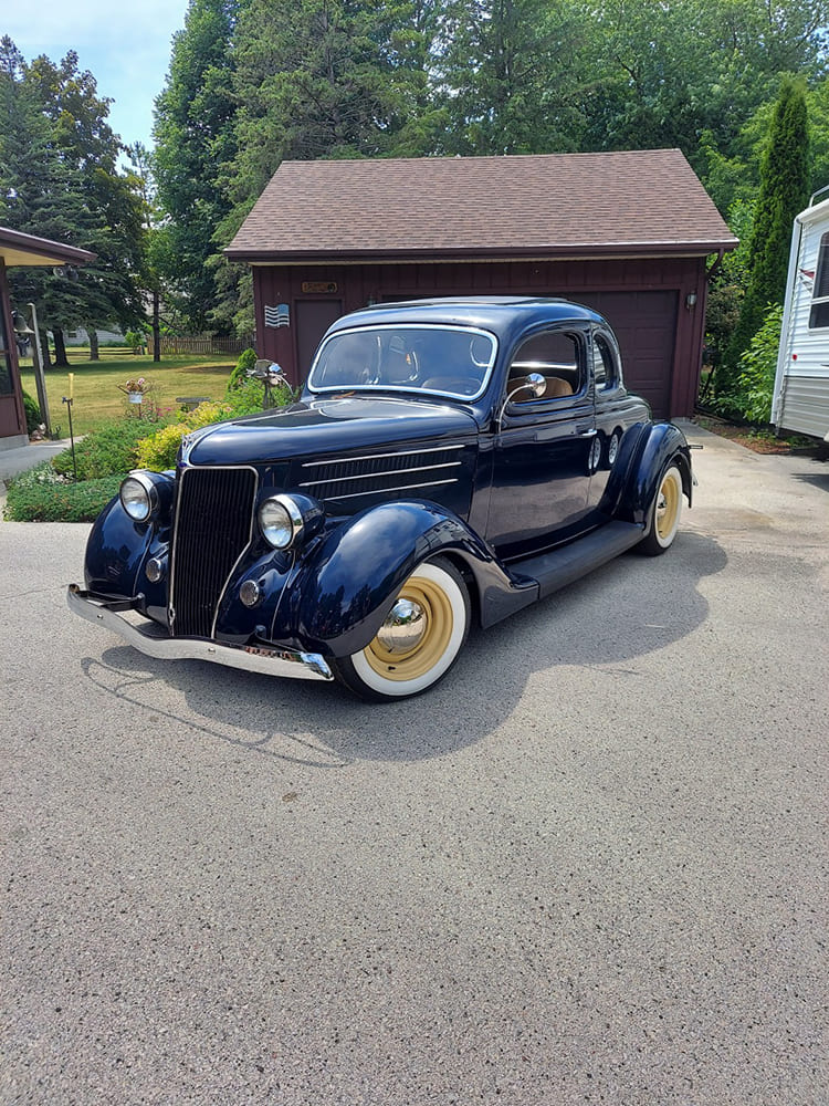 Low front three-quarter perspective of a glossy midnight blue 1936 Ford street rod coupe featuring cream-colored steel wheels with wide whitewall tires.