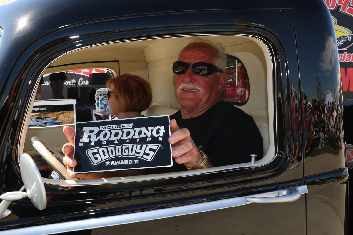 Driver with mustache and sunglasses seated in a classic black hot rod truck holding out his Modern Rodding Magazine Goodguys Award plaque.