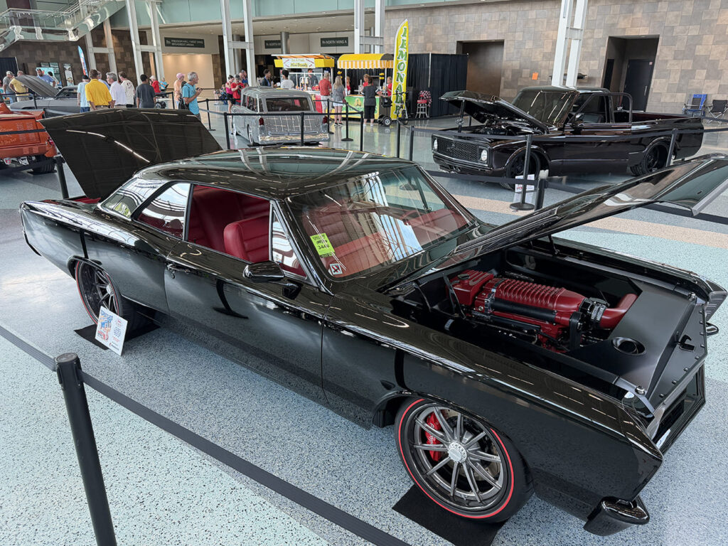 High front three-quarter view of a jet-black 1966 Plymouth Belvedere muscle car featuring an open hood and trunk, red leather upholstery, red supercharger, and redline tires.