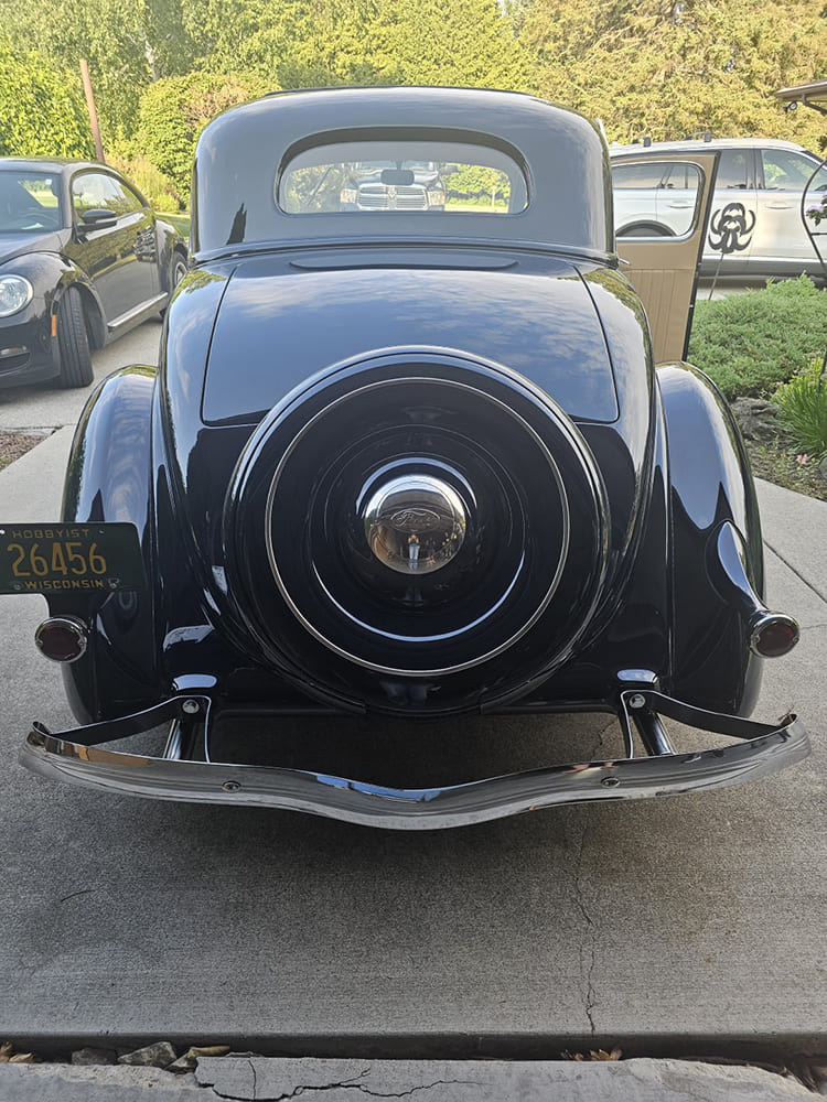 Direct rear view of a dark blue 1936 Ford coupe showing a rear-mounted enclosed spare tire, curved chrome bumper, and classic Wisconsin hobbyist license plate.