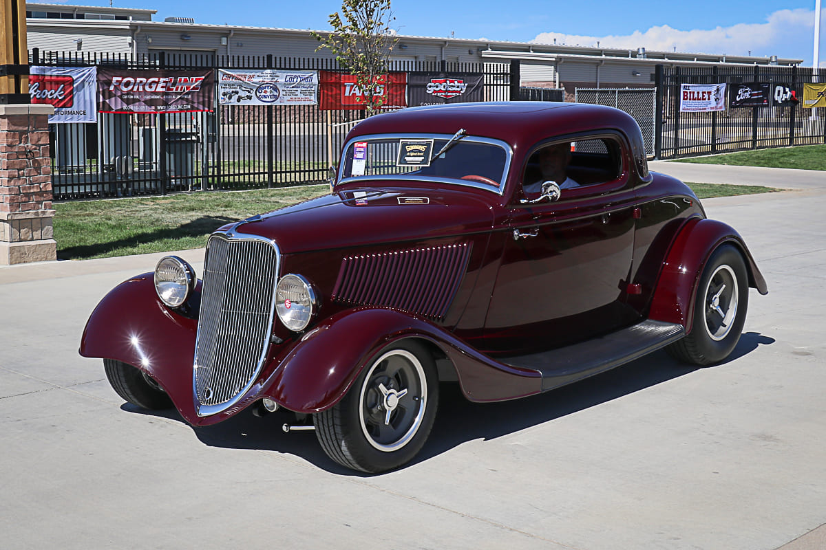 Front three-quarter shot of a deep metallic maroon 1934 Ford coupe street rod driving past sponsor fence banners under a blue sky.