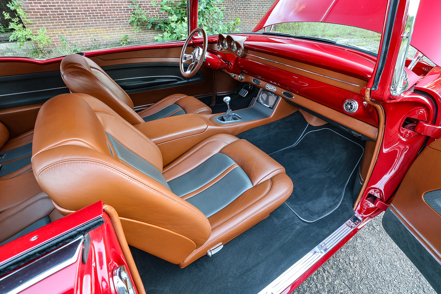 Driver-side cabin view of a red 1955 Bel Air showcasing a painted dashboard, aftermarket gauges, floor shifter, and tan leather seats.