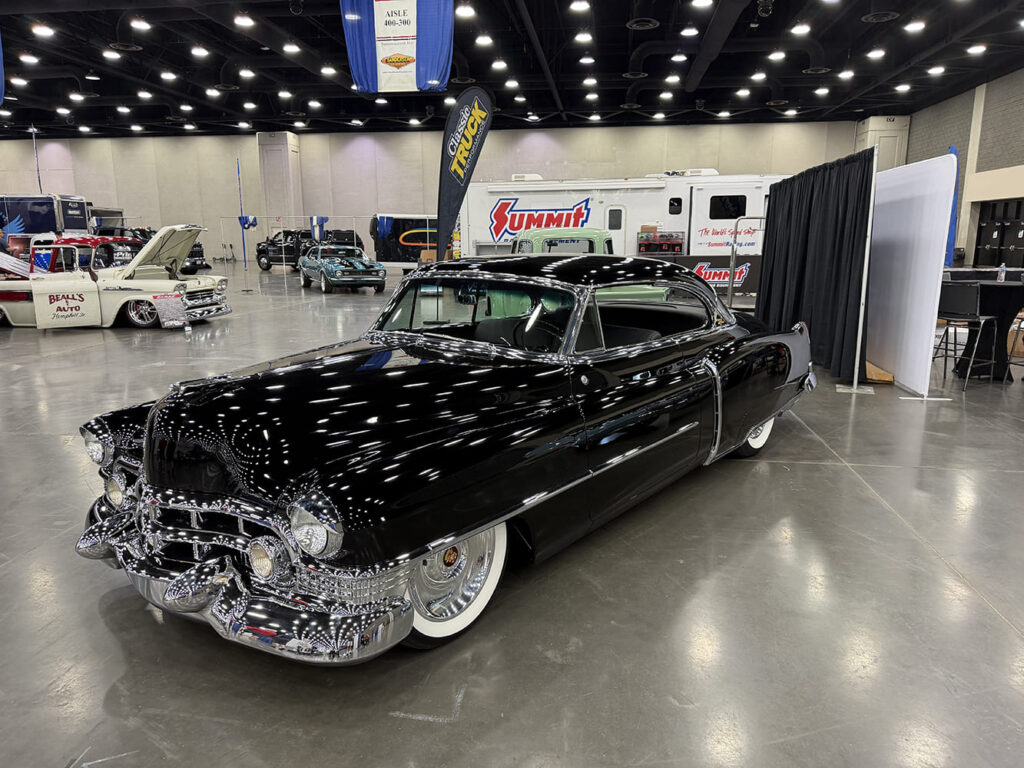 Low-angle front three-quarter shot of a mirror-finish black 1954 Cadillac coupe with heavy chrome grille, chrome hubcaps, and wide whitewall tires on an exhibition hall floor.