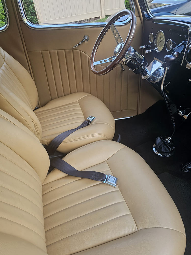Driver-side interior view of a 1936 Ford coupe highlighting pleated tan leather bucket seats, lap belts, floor shifter, and wood-rimmed steering wheel.