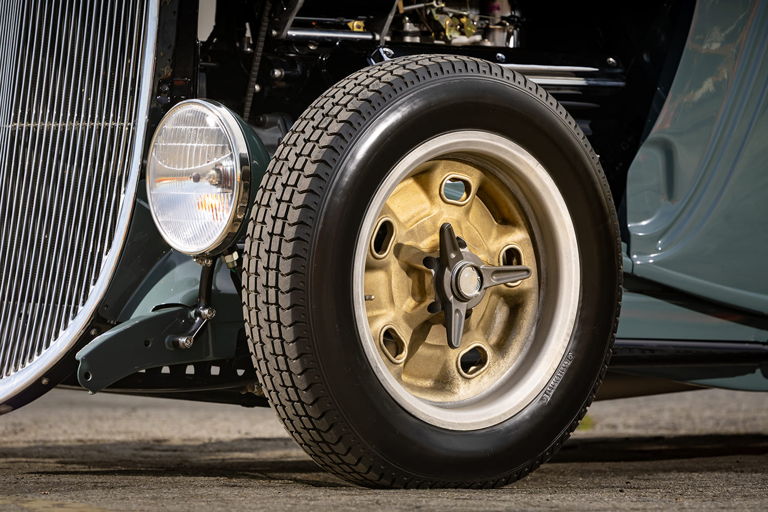 Detail of the front chrome headlight, exposed axle, and vintage gold wheel with a three-ear knock-off spinner on a 1933 Ford hot rod.