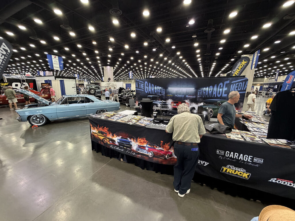 Attendees and staff browsing print magazines and merchandise at the In The Garage Media expo booth, with a light blue Chevrolet Chevelle displayed inside the convention hall.