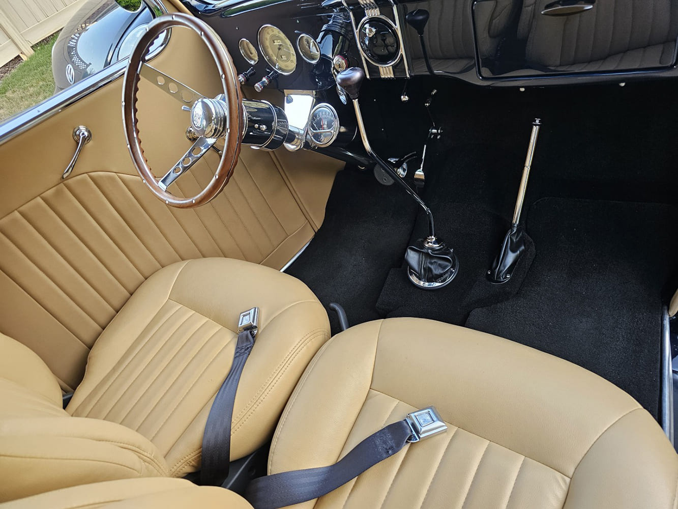 High-angle cockpit view of a 1936 Ford street rod showing the painted gloss black dashboard, original-style round gauge cluster, tall floor shifter, and tan upholstery.