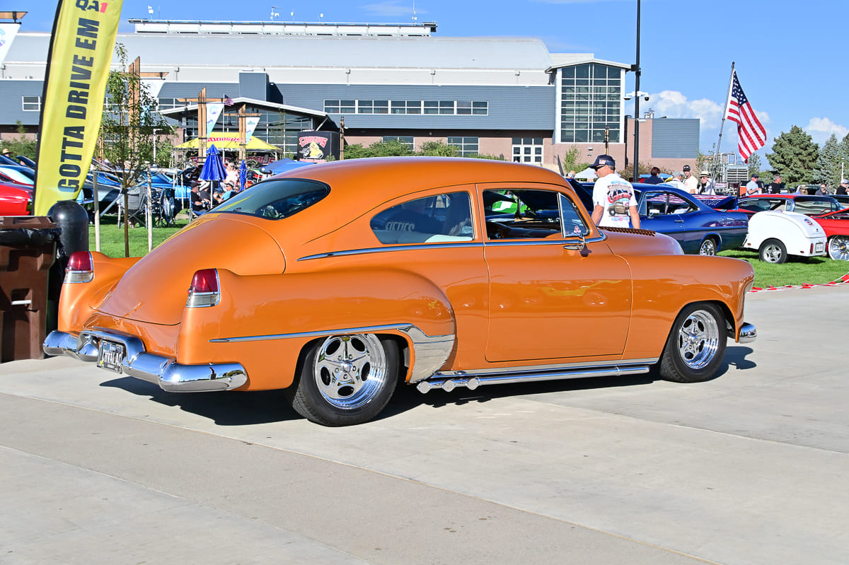 Rear three-quarter view of a lowered metallic copper-orange classic fastback coupe with side lake pipes parked near event flags.