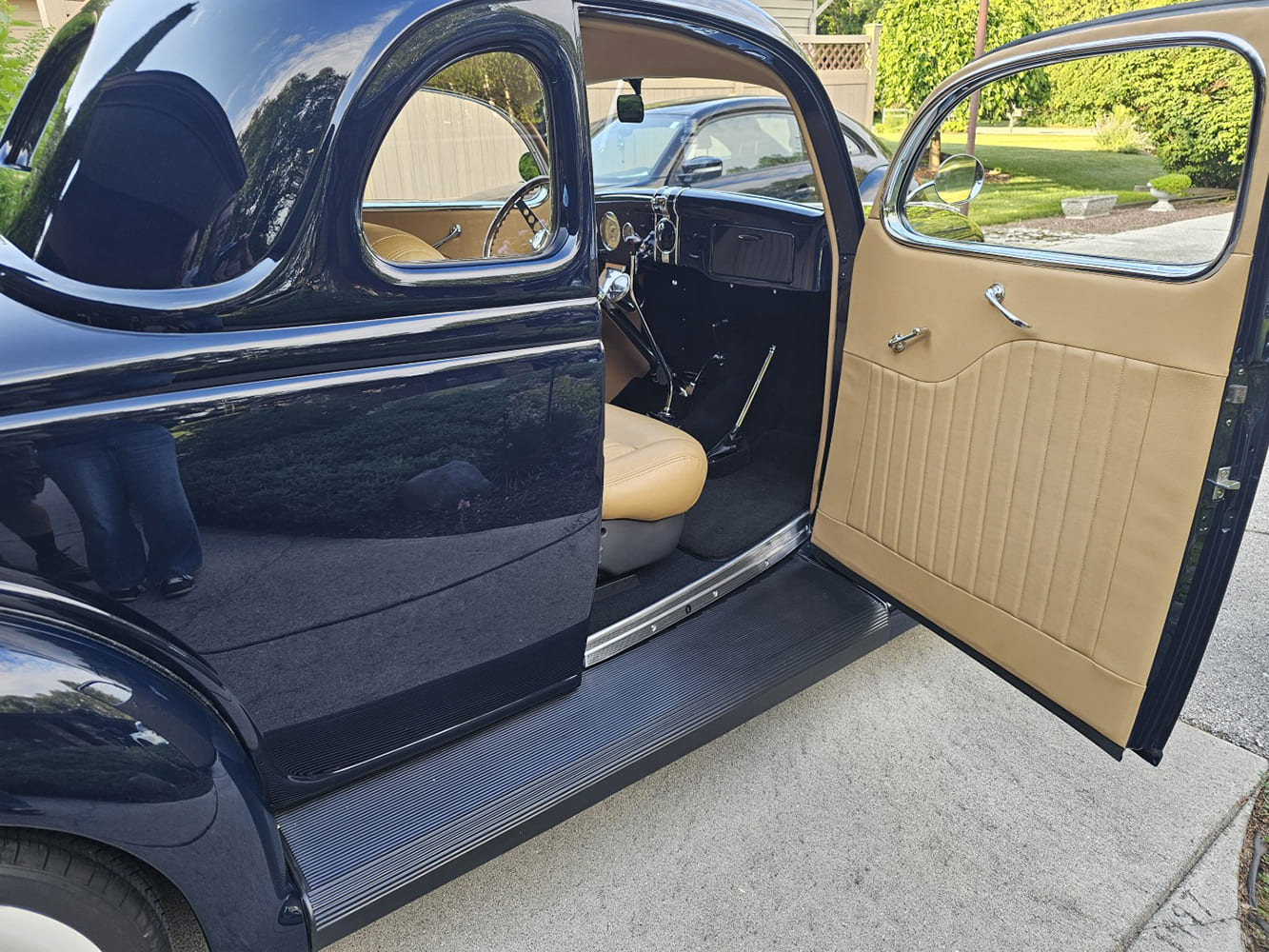 Looking through the open passenger door of a dark blue 1936 Ford coupe showing tan leather door cards, running board, and interior bucket seats.