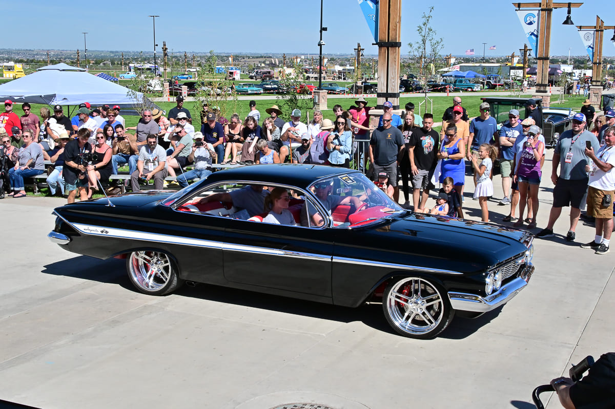 Black 1961 Chevrolet Impala bubbletop coupe with red interior and chrome multi-piece wheels cruising past an outdoor crowd