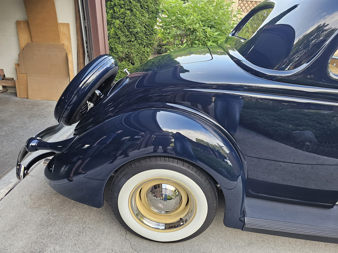 Low-angle rear quarter view of a midnight blue 1936 Ford coupe showing the rear-mounted spare, rounded fender, and cream wheel with wide whitewall tire.