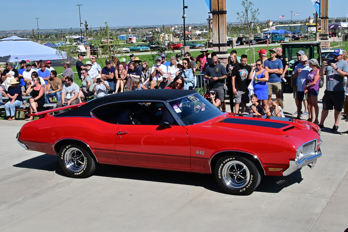 Front three-quarter angle of a red 1970 Oldsmobile 442 hardtop with black hood stripes and Cooper Cobra tires driving past spectators at Good Guys Colorado.
