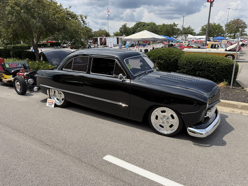 Low-angle front three-quarter view of a lowered jet-black Ford Shoebox coupe featuring a louvered hood, custom grille teeth, and polished wheels on asphalt.