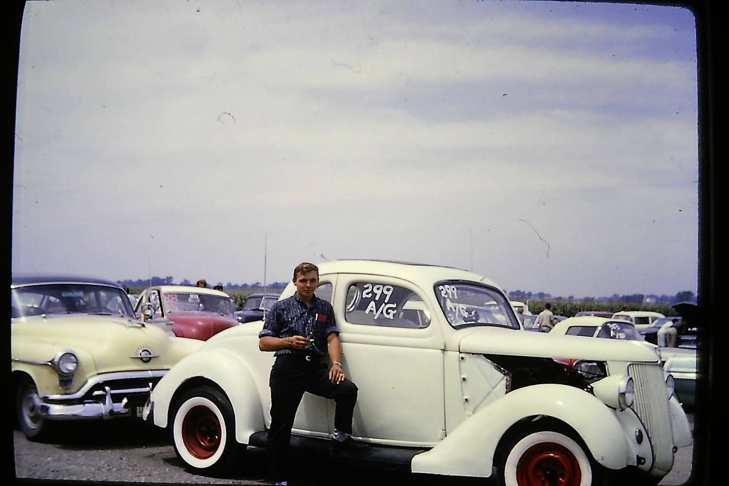 Historic color photograph of a young man leaning against a white 1936 Ford A/Gas drag car with red wheels and whitewalls in the dragway pits.