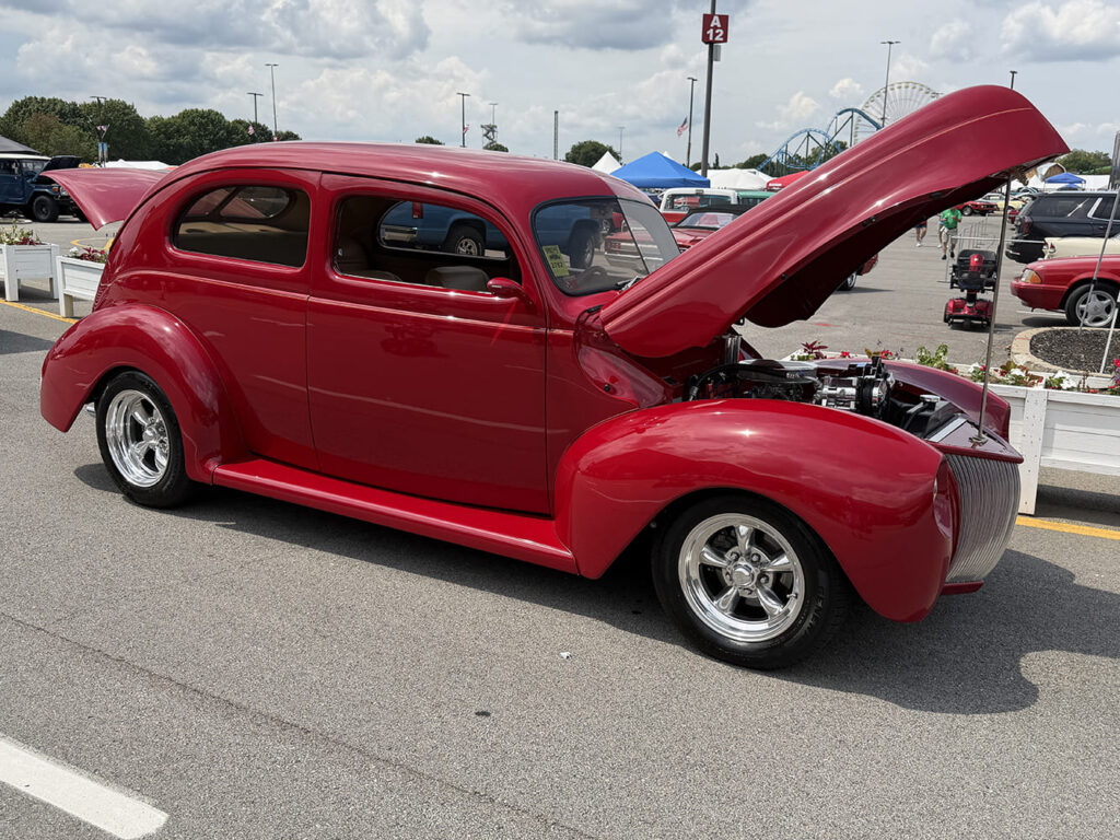 Front three-quarter angle of a deep cherry red 1940 Ford deluxe coupe street rod with an open hood displaying a chrome-detailed V8 engine in the show lot.