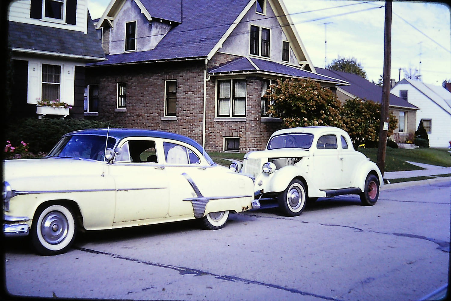 Vintage street photo of a two-tone 1950s passenger car flat-towing a white 1936 Ford coupe hot rod on a residential street.