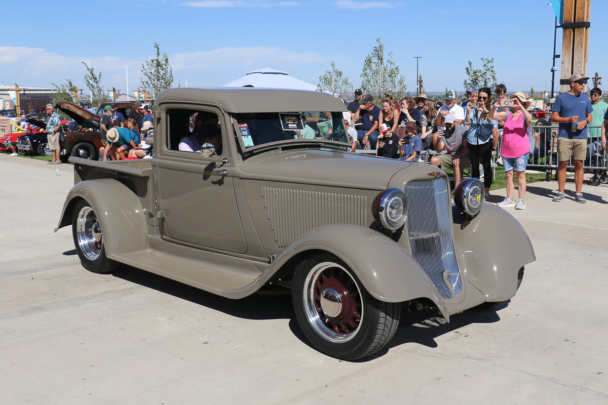 Front three-quarter view of a taupe gray 1934 Ford pickup hot rod with custom maroon wire-style artillery wheels rolling past car show crowds.