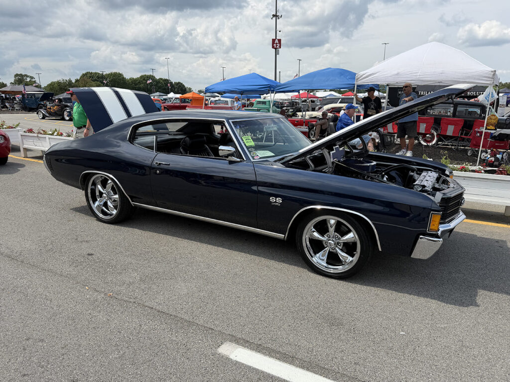 Driver-side profile of a midnight blue Chevrolet Chevelle SS with white racing stripes, open hood and trunk, and chrome multi-spoke wheels on the showgrounds.