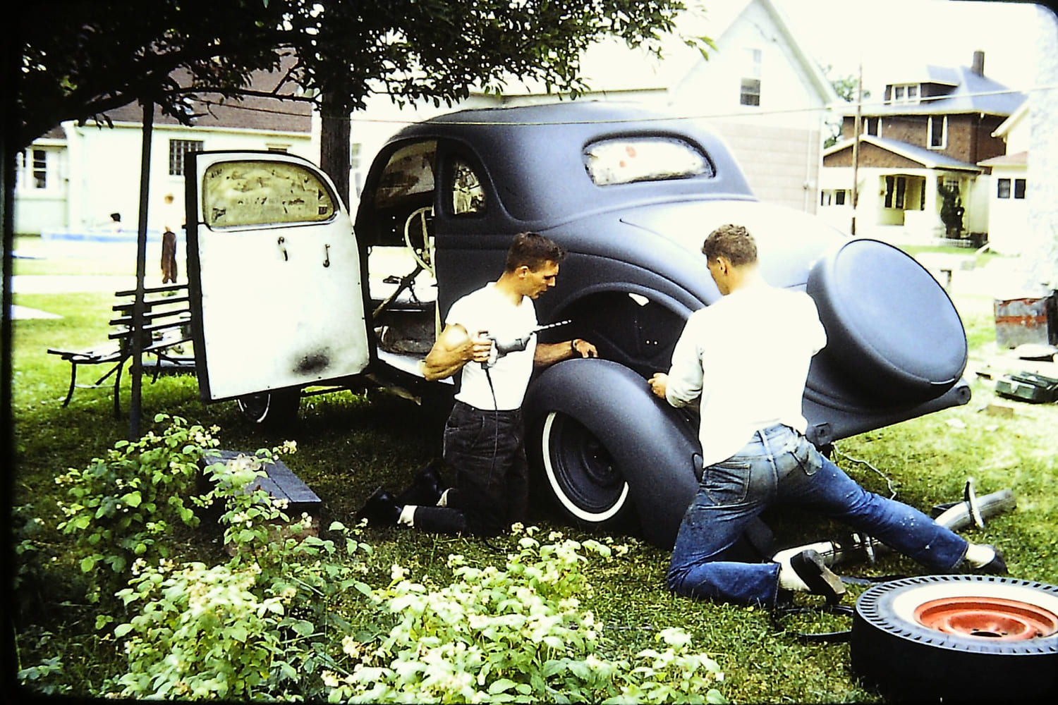 Vintage color photo of two young men working on the rear fender and primer-coated body of a 1936 Ford coupe outdoors on the grass.