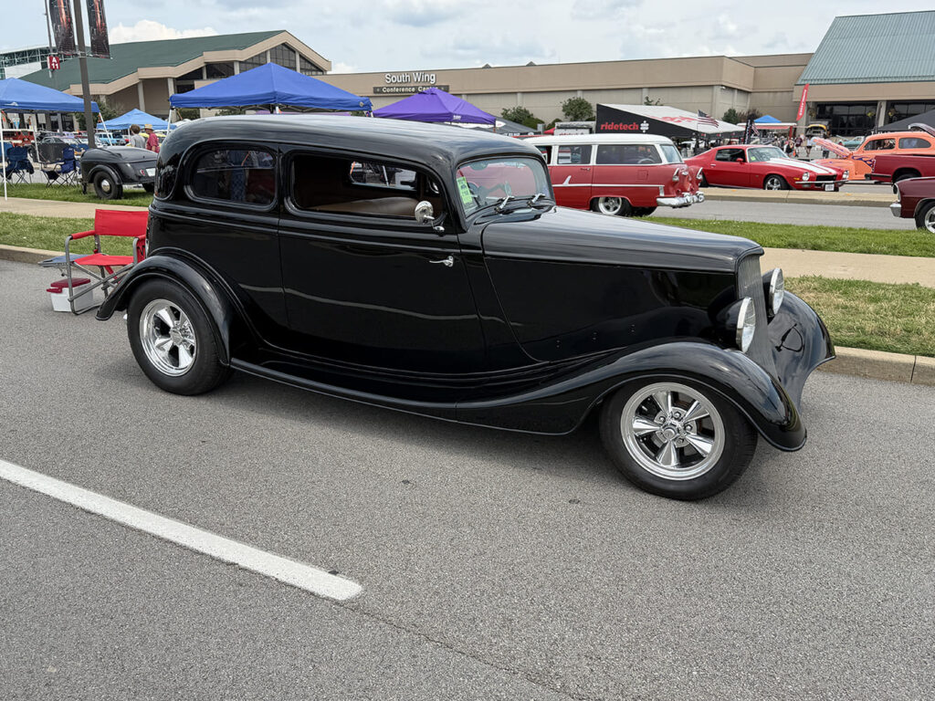 Front three-quarter perspective of a high-gloss black 1933 Ford two-door sedan street rod with polished five-spoke wheels parked beside outdoor vendor tents.
