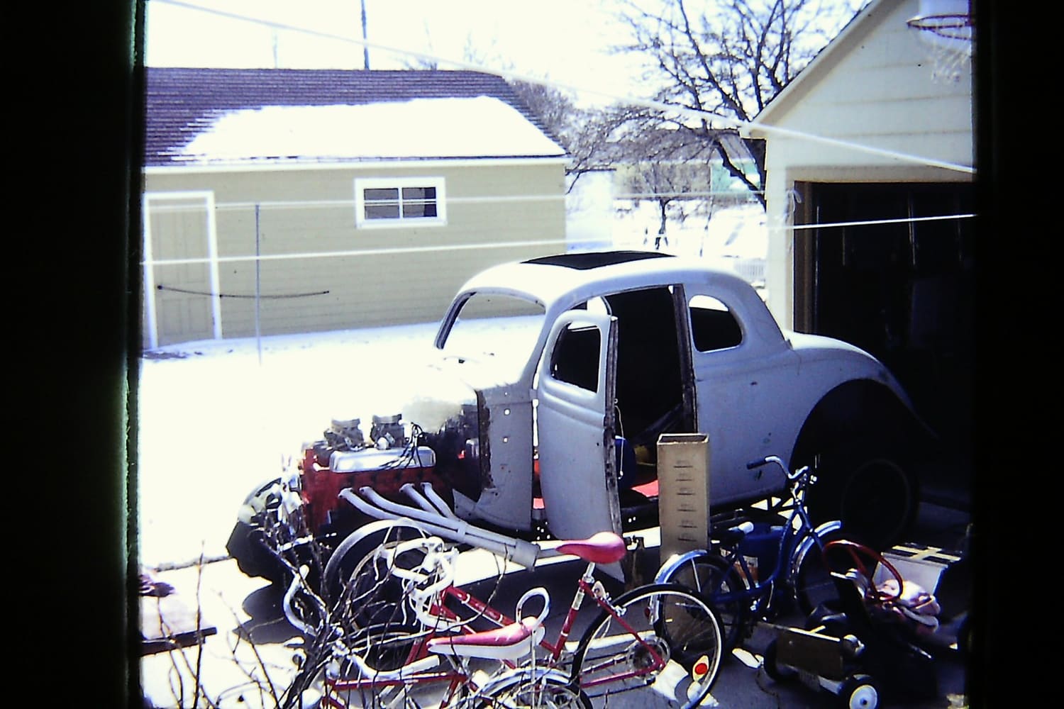 Vintage view from a window showing a primer-gray 1936 Ford coupe project car with an exposed dual-carb V8 and white headers parked in the snow.