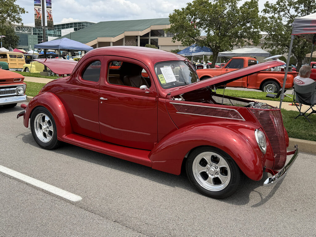 High front three-quarter view of a metallic crimson 1937 Ford five-window coupe with an open hood, fine-tooth grille, and polished billet wheels.
