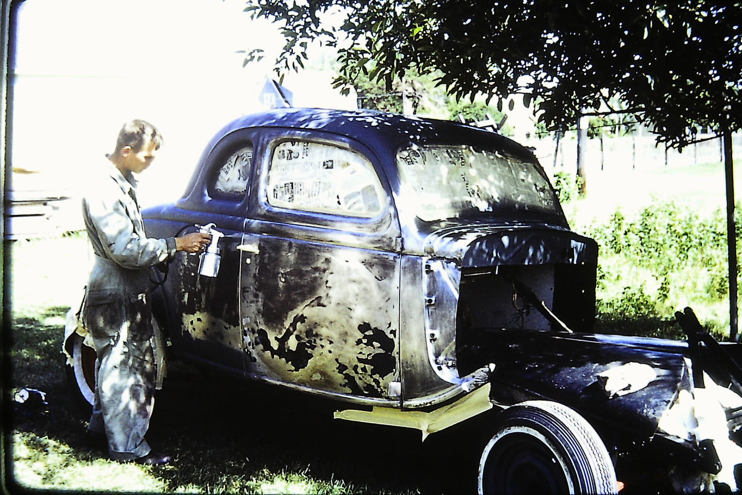 Vintage photograph of a man in coveralls using a handheld spray gun to paint the stripped metal and primer body of a 1936 Ford coupe under trees.