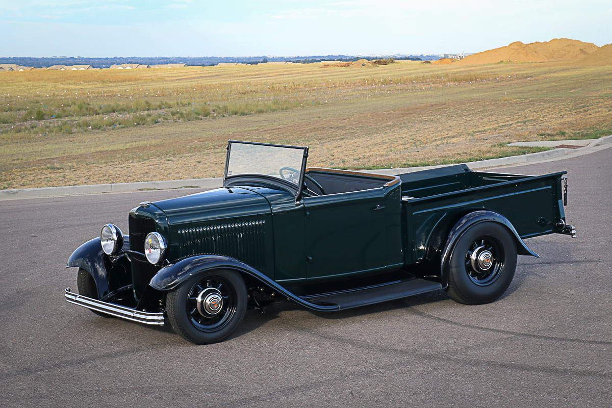 High front three-quarter perspective of a fendered dark green 1932 Ford roadster pickup street rod with black steel wheels parked along an asphalt road.