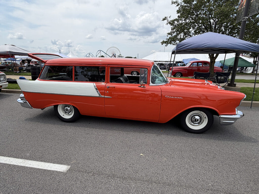 Direct side profile of a two-tone coral orange and white 1957 Chevrolet two-door station wagon with white steel wheels and dog dish hubcaps parked under a canopy.