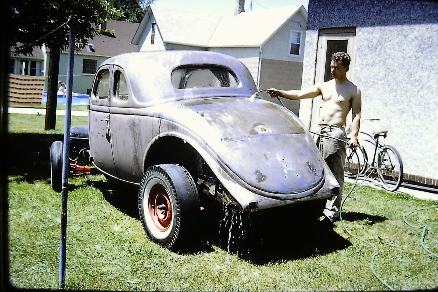 Historic photo of a shirtless man spraying water onto the stripped steel body and rear deck of a 1936 Ford coupe hot rod in a backyard.