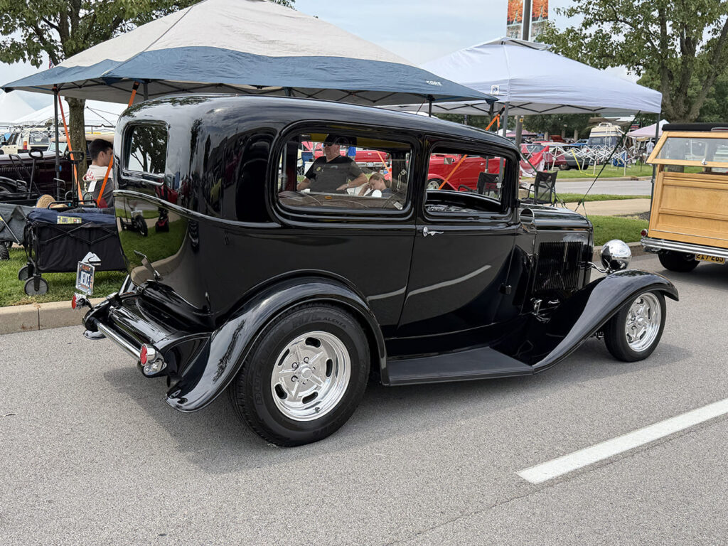 Rear three-quarter view of a glossy black 1932 Ford Tudor sedan street rod featuring polished wheels, curved rear fenders, and dual chrome exhaust tips outdoors.