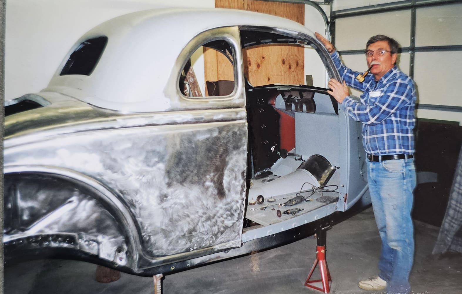 Tom Lawrence with glasses and a pipe smiling while standing beside his stripped 1936 Ford coupe body mounted on jack stands inside a home garage.