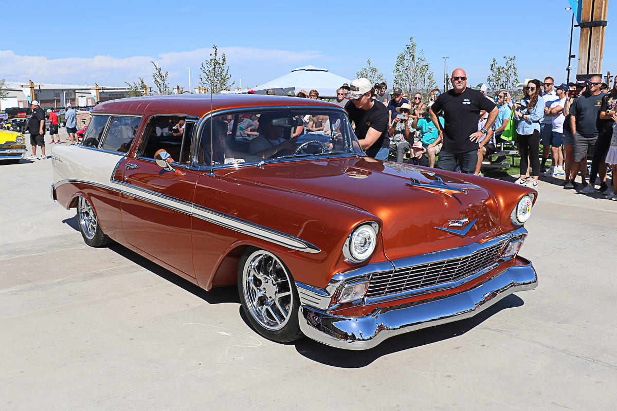 Front three-quarter view of a customized metallic copper-brown and cream 1956 Chevrolet Nomad wagon driving through crowds at the Colorado Nationals car show.