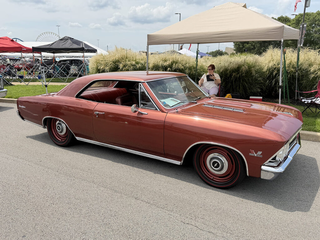 Front three-quarter angle of a lowered metallic copper-bronze 1966 Chevrolet Chevelle hardtop with color-matched steel wheels and redline tires parked under a canopy.