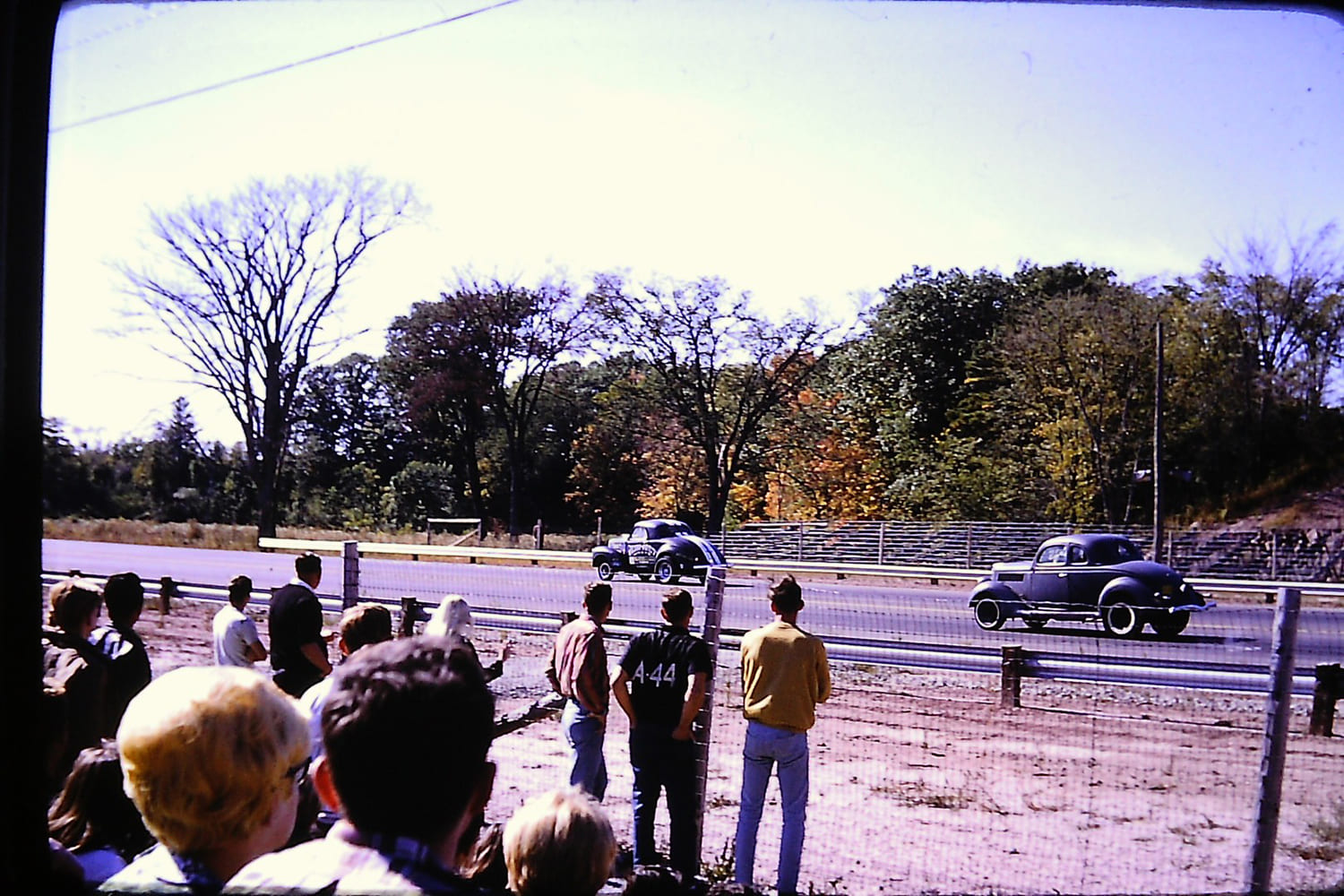 Historic color photograph from behind a spectator chain-link fence showing two vintage 1936 Ford coupes racing side by side down an asphalt drag strip.