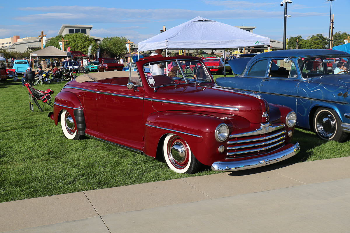 Low front three-quarter profile of a lowered maroon 1947 Ford convertible custom with whitewall tires, chrome hubcaps, and tan soft top parked on grass.